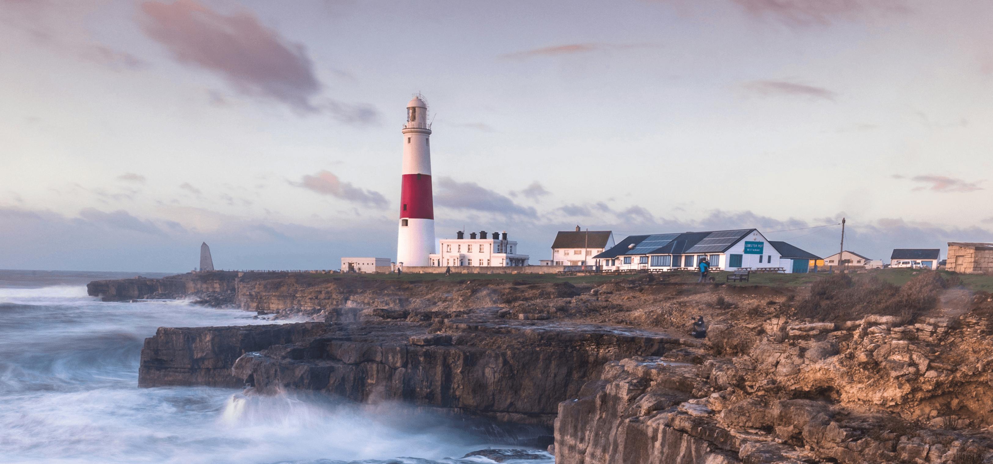 Le onde si infrangono su una scogliera britannica con un faro in lontananza