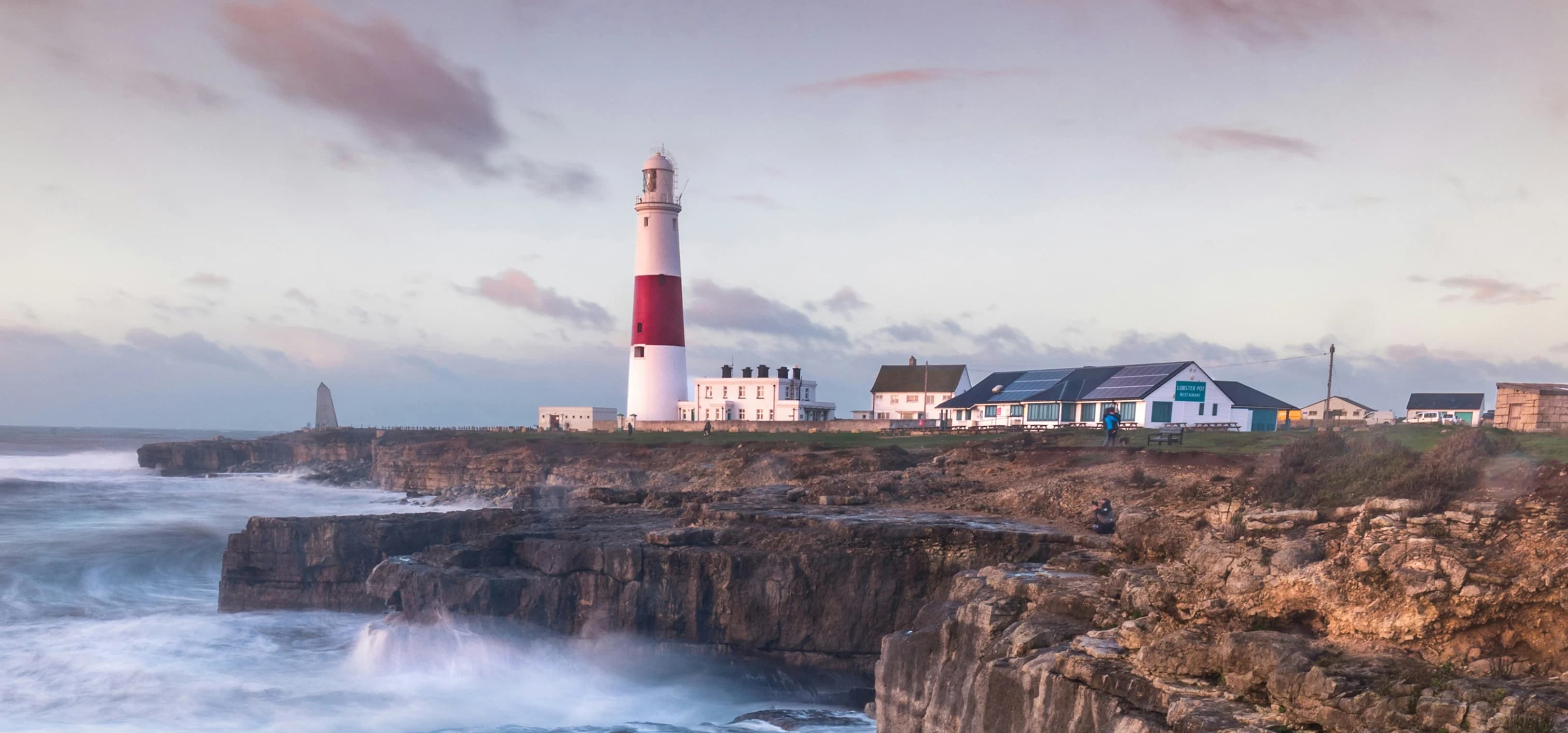 Le onde si infrangono su una scogliera britannica con un faro in lontananza