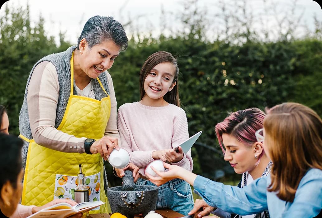 Un adulto insegna a un gruppo di bambini a preparare degli ingredienti utilizzando un mortaio e un pestello durante un'attività pratica all'aperto dedicata alla scoperta di una cultura.