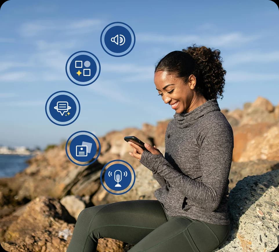 Donna seduta all'aperto su alcune rocce vicino all'acqua mentre sorride e guarda il telefono. Attorno a lei sono visibili le icone delle funzionalità di Rosetta Stone Sapphire.